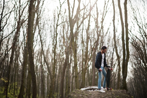 Hombre árabe de estilo callejero en anteojos con longboard en madera.