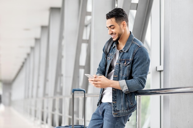 Hombre árabe feliz enviando mensajes en el teléfono inteligente mientras espera con equipaje en el aeropuerto
