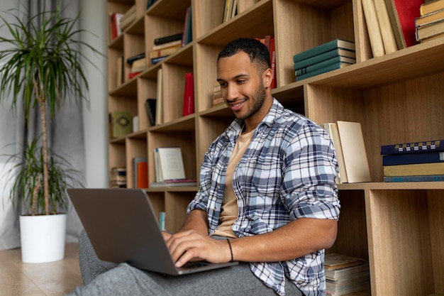 Hombre árabe feliz escribiendo en el teclado de la computadora portátil mientras se apoya en la estantería y estudia o trabaja desde casa