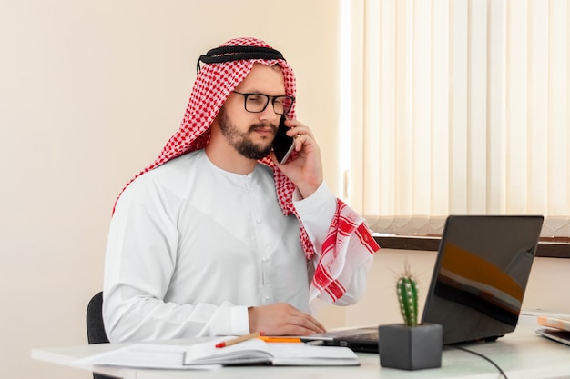 Un hombre árabe, un hombre de negocios, un jeque con un traje nacional árabe está hablando por un teléfono inteligente en una mesa de la oficina. Inversiones, negocios, trabajo vía Internet, contratos online.