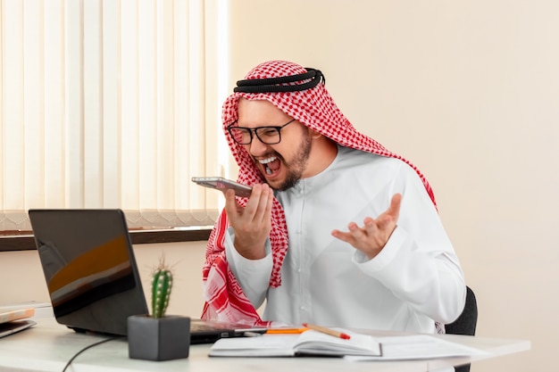 Un hombre árabe, un hombre de negocios, un jeque con un traje nacional árabe le grita mal a alguien durante una conversación. Inversiones, negocios, trabajo vía Internet, contratos online.