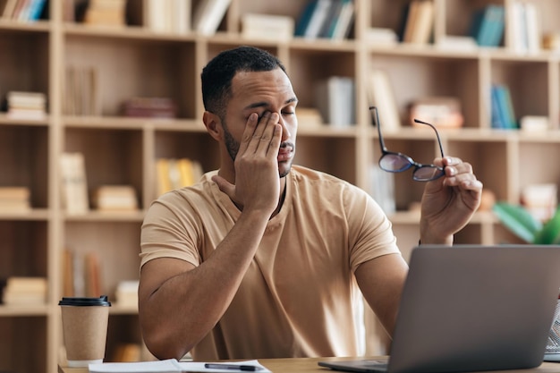 Hombre árabe independiente cansado que trabaja en una computadora portátil desde la oficina en casa y se quita las gafas sentado en el escritorio