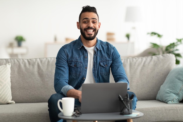 Hombre árabe sentado en un sofá con una computadora portátil participando en conferencias web o videoconferencias trabajando o estudiando en línea