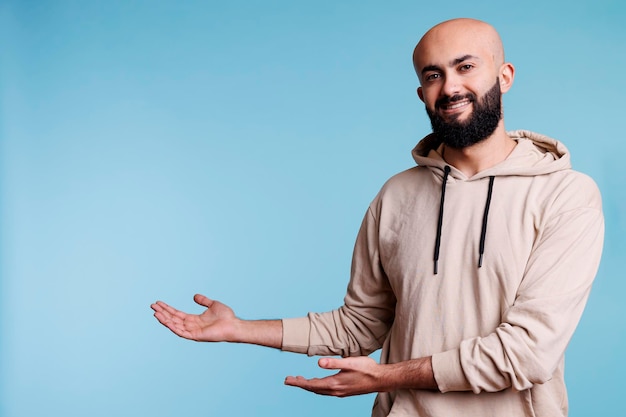 Un hombre árabe sonriente y despreocupado que apunta al retrato de estudio en la dirección correcta. Joven promocionando productos, posando mientras muestra a un lado y mirando a la cámara con expresión alegre