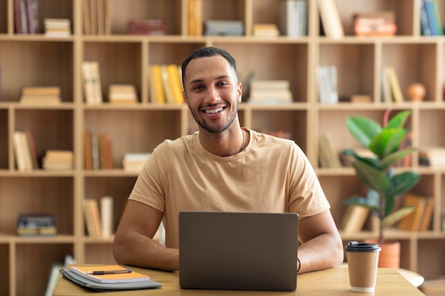 Hombre árabe sonriente sentado en el escritorio trabajando en una computadora portátil en el interior en el espacio de copia de la sala de estar Concepto de trabajo distante