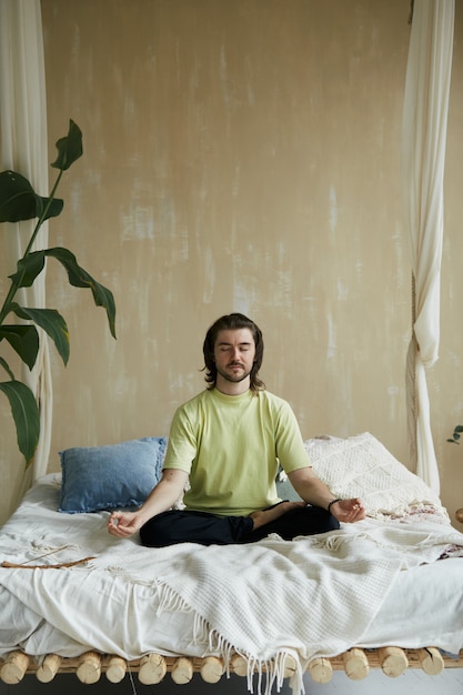 Hombre armónico en postura de loto en la cama, persona relajada en asanas de yoga y mudra practicando meditación en casa en la cama