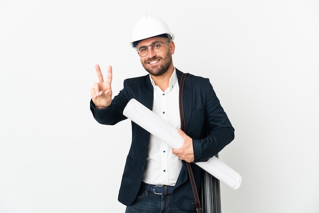 Hombre arquitecto con casco y sosteniendo planos aislados en blanco sonriendo y mostrando el signo de la victoria