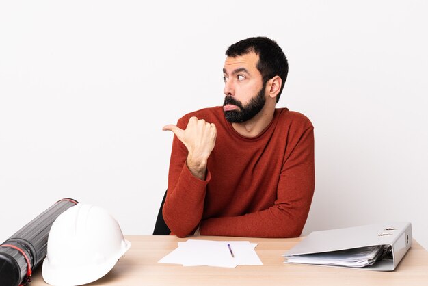 Hombre de arquitecto caucásico con barba en una mesa infeliz y apuntando hacia un lado.