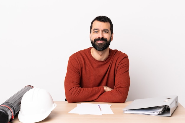 Hombre arquitecto caucásico con barba en una mesa manteniendo los brazos cruzados en posición frontal.