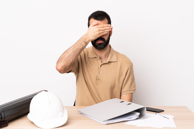 Hombre de arquitecto caucásico con barba en una mesa que cubre los ojos con las manos