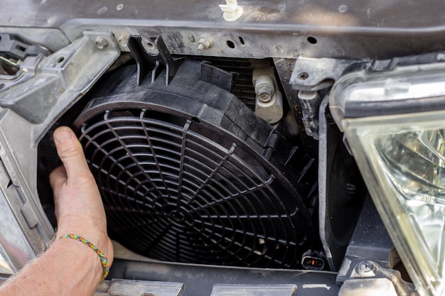 Un hombre arregla un ventilador de refrigeración del motor debajo de la parrilla del radiador de un automóvil Mantenimiento y reparación de máquinas