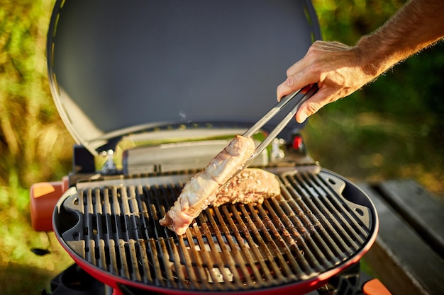 Hombre asado carne en la parrilla de gas en la parrilla de barbacoa al aire libre en el patio trasero, carne de bistec a la parrilla, picnic familiar de verano, comida en la naturaleza.