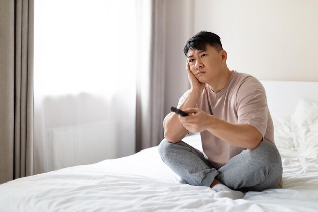 Hombre asiático aburrido sentado en la cama en los canales de casa en la televisión