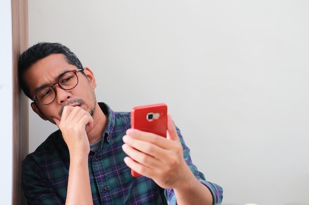 Hombre asiático adulto apoyado en la pared que muestra una expresión triste al mirar su teléfono