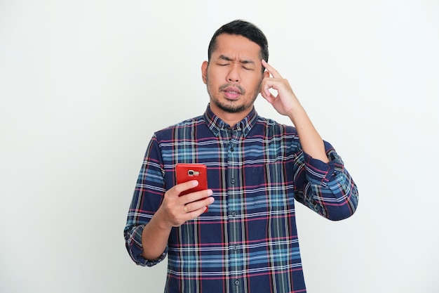 Foto hombre asiático adulto intenta recordar algo con los ojos cerrados mientras sostiene el teléfono móvil