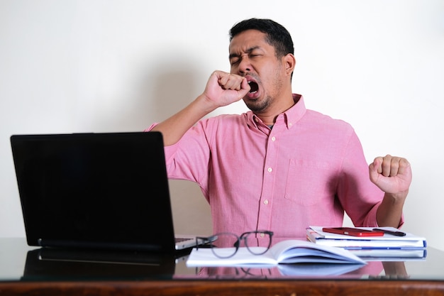 Hombre asiático adulto mostrando gesto cansado frente a su computadora portátil de trabajo