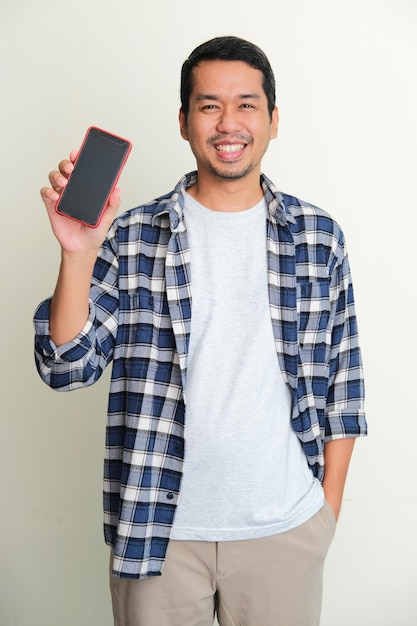 Hombre asiático adulto de pie mientras sonríe mostrando la pantalla del teléfono móvil en blanco que sostiene