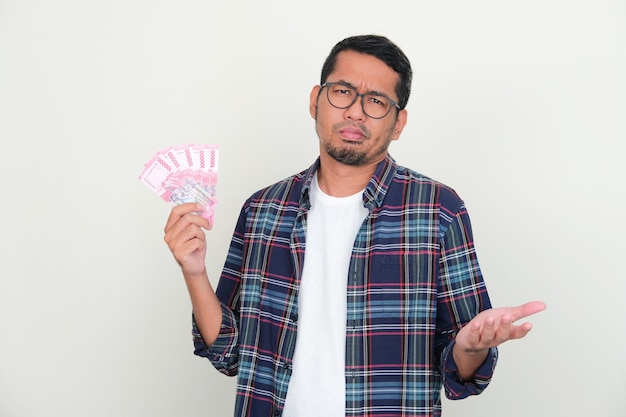 Hombre asiático adulto que muestra una expresión confusa mientras sostiene dinero