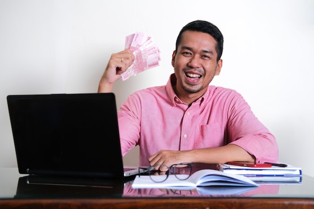 Hombre asiático adulto que muestra la expresión feliz mientras sostiene el papel moneda en su mesa de trabajo