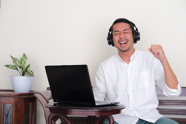 Hombre asiático adulto que muestra felicidad al escuchar música y sentarse frente a la computadora portátil