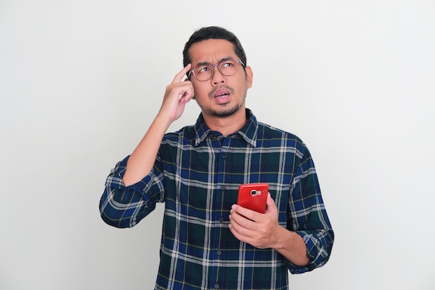 Hombre asiático adulto que muestra una pose de pensamiento mientras sostiene su teléfono