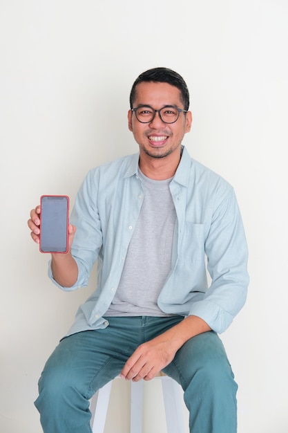 Hombre asiático adulto sentado en un banco que muestra la pantalla del teléfono en blanco con expresión feliz