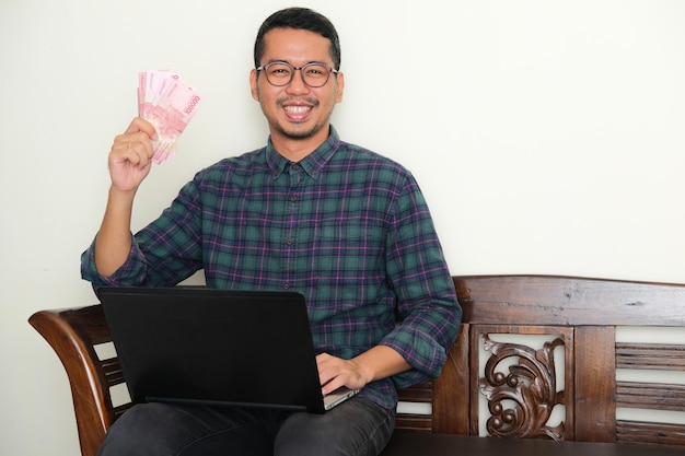 Hombre asiático adulto sentado en un banco sosteniendo papel moneda y mostrando expresión feliz