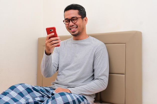 Hombre asiático adulto sentado en la cama sonriendo feliz mientras mira su teléfono móvil