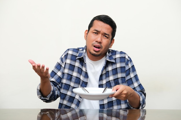 Hombre asiático adulto sentado en la mesa del comedor que muestra una expresión confusa mientras muestra un plato vacío