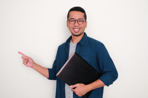 Hombre asiático adulto sonriendo y apuntando hacia el lado derecho mientras sostiene una computadora portátil