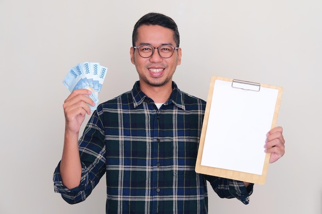 Hombre asiático adulto sonriendo confiado mientras sostiene dinero y papel blanco en blanco en el portapapeles