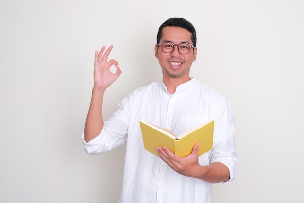 Hombre asiático adulto sonriendo y dando la señal de OK con el dedo mientras sostiene un libro