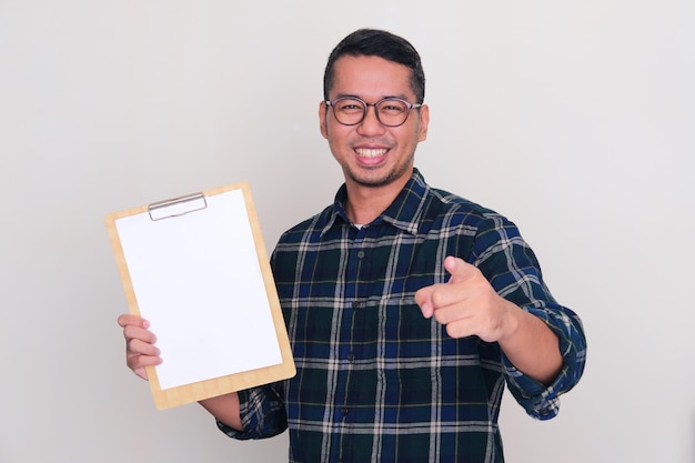 Hombre asiático adulto sonriendo emocionado y señalando a alguien mientras sostiene un papel blanco vacío en el portapapeles