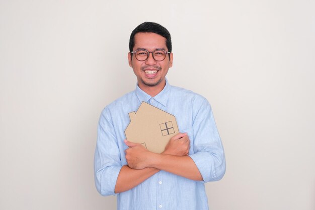 Hombre asiático adulto sonriendo feliz al abrazar un cartón con forma de casa
