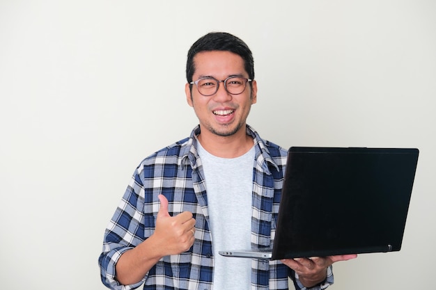 Hombre asiático adulto sonriendo feliz y dando pulgar hacia arriba mientras sostiene una computadora portátil