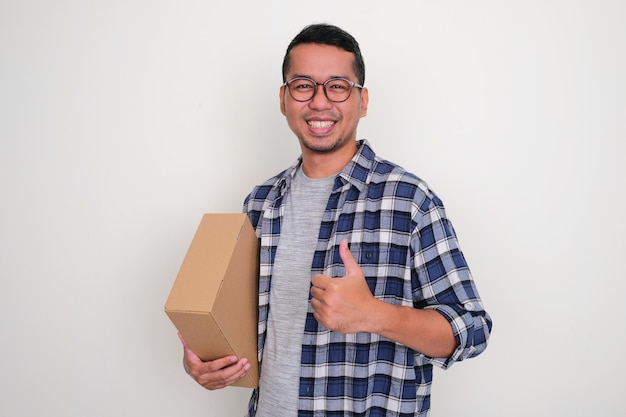 Hombre asiático adulto sonriendo feliz y levantando el pulgar cuando sostiene una caja de paquetes