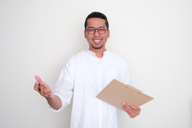 Foto hombre asiático adulto sonriendo haciendo pose de saludo mientras sostiene un bolígrafo y un portapapeles