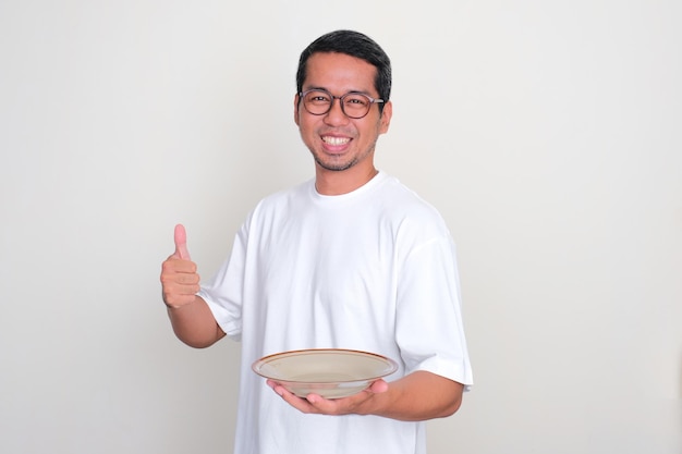 Hombre asiático adulto sonriendo y levantando el pulgar mientras muestra un plato de comedor vacío