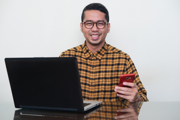 Hombre asiático adulto sonriendo mientras se sienta frente a la computadora portátil y sostiene el teléfono