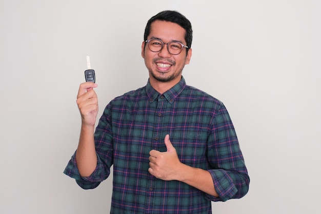 Foto hombre asiático adulto sonriendo orgulloso y levantando el pulgar mientras sostiene una llave de auto