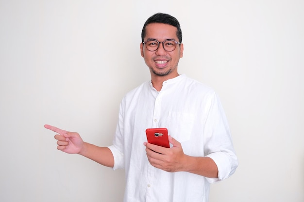 Hombre asiático adulto sonriendo y señalando hacia el lado derecho mientras sostiene su teléfono