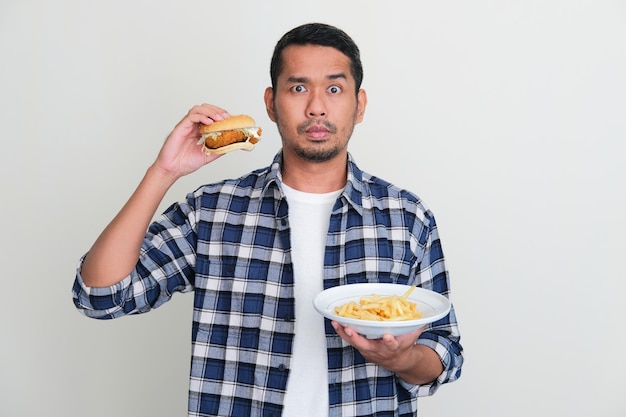 Hombre asiático adulto sosteniendo una hamburguesa y papas fritas con expresión seria