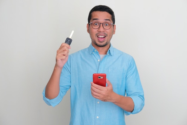 Hombre asiático adulto sosteniendo la llave del auto y el teléfono con expresión asombrada