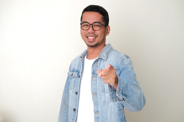 Hombre asiático adulto vestido con chaqueta de jeans azul apuntando hacia adelante mientras sonríe feliz