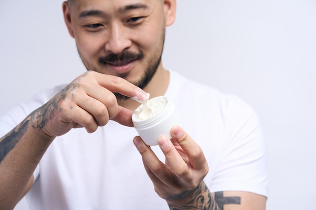 Hombre asiático alegre aplicando crema en la cara con frasco humectante en la mano