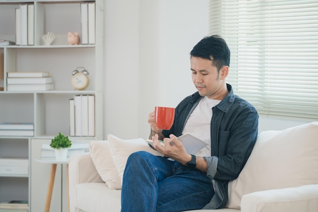 Un hombre asiático alegre leyendo un libro y tomando café caliente mientras está sentado en el sofá de la sala de estar en casa Un hombre asiático relex leyendo un libro sonríe en el sofá En la casa Actividad pasatiempo en el concepto de casa