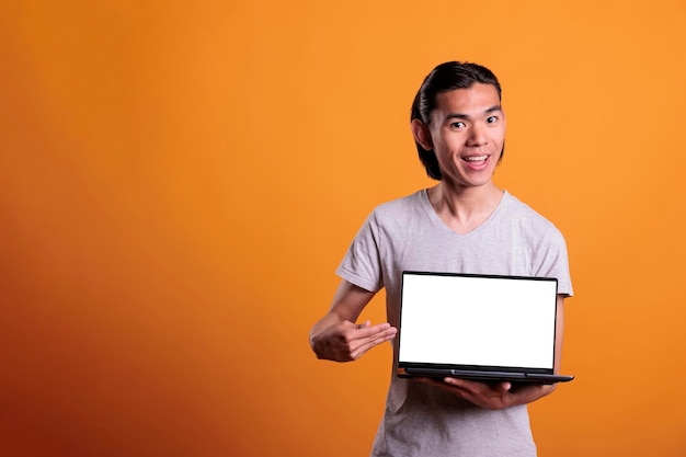 Hombre asiático alegre que anuncia portátil con pantalla blanca en blanco, maqueta de promoción de software con espacio de copia. Adolescente sonriente apuntando a un ordenador portátil con pantalla vacía