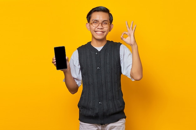 Hombre asiático alegre que muestra la pantalla en blanco del teléfono inteligente y gesticula bien firmar sobre fondo amarillo