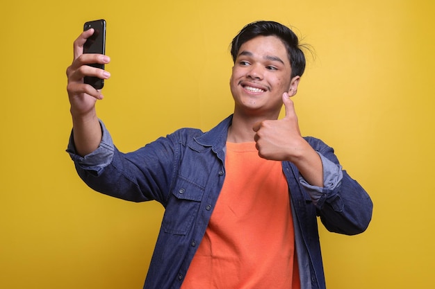 Hombre asiático alegre tomando una selfie y mostrando el pulgar hacia arriba en un fondo amarillo