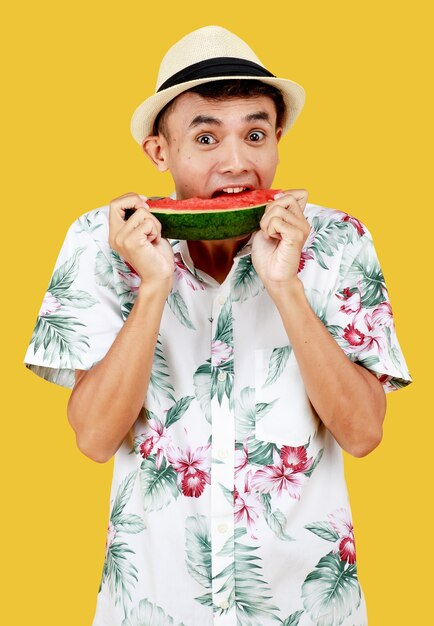 Hombre asiático atractivo joven en camisa blanca hawaiana con sombrero blanco comiendo una rebanada de sandía con entusiasmo sobre fondo amarillo. Concepto de vacaciones en la playa.